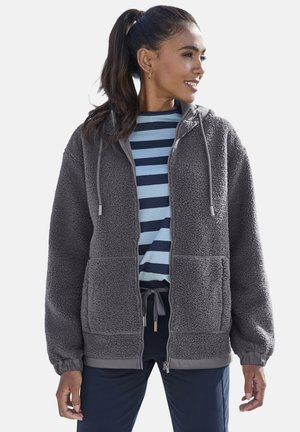 Frau mit dunklen Haaren im Pferdeschwanz, die eine graue Fleecejacke mit Reißverschluss über einem blau gestreiften Hemd und dunkler Hose trägt und nach rechts schaut.