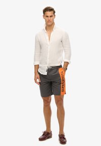 Grijze zwembroek met een oranje accent waarop "SUPERDRY" in het zwart staat. Het model draagt een wit linnen overhemd en bruine boot schoenen.