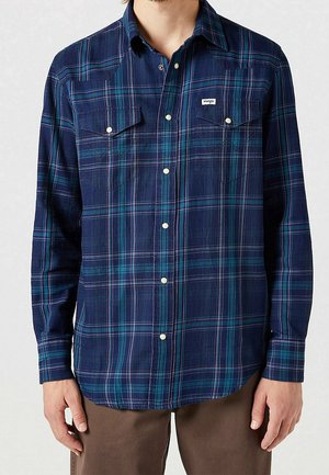 Mann trägt ein blau-grünes kariertes Hemd mit langen Ärmeln und Brusttaschen über einer braunen Hose.