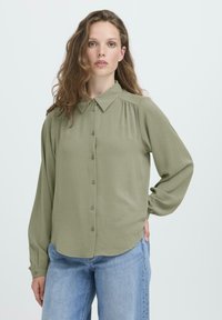 Sieviete ar gariem viļņainiem matiem, tērpusies brīvā olīvzaļās krāsas krekliņā ar pogām un gaiši zilos džinsos, stāvot ar vienu roku kabatā.