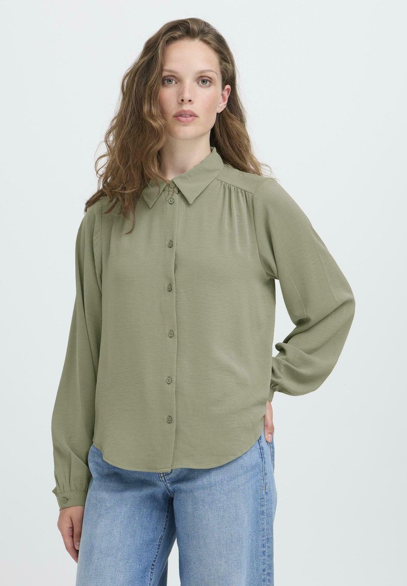 Sieviete ar gariem viļņainiem matiem, tērpusies brīvā olīvzaļās krāsas krekliņā ar pogām un gaiši zilos džinsos, stāvot ar vienu roku kabatā.