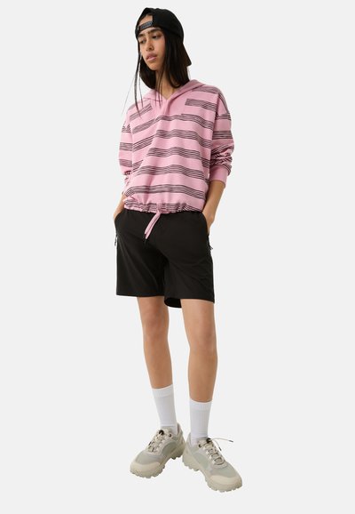 Helle rosa Kapuzenjacke mit schwarzen Streifen, Kordelzugbund, kombiniert mit schwarzen Shorts und klobigen beigen Turnschuhen. Das Modell trägt weiße Socken und eine Kappe.