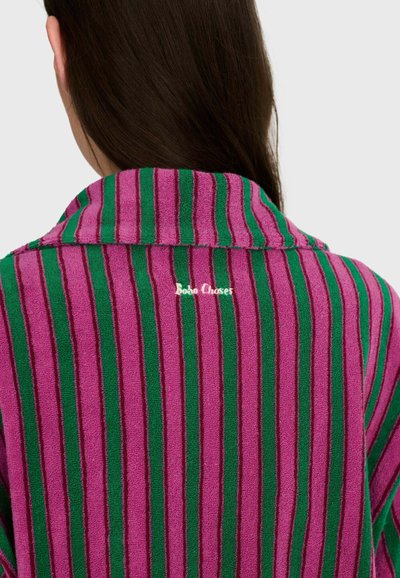 Femme aux longs cheveux bruns portant une chemise en éponge rayée verticalement violet et vert avec col, vue de dos.