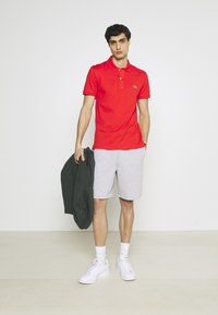 Rode poloshirt met een kraag, grijze shorts, witte sneakers en een grijze hoodie over de arm gedrapeerd. Model staat tegen een effen achtergrond.
