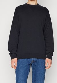 Schwarzer Rundhals-Sweatshirt mit gerippten Bündchen und Saum, ausgestattet mit einem strukturierenden Strickmuster. Kombiniert mit blauen Jeans.