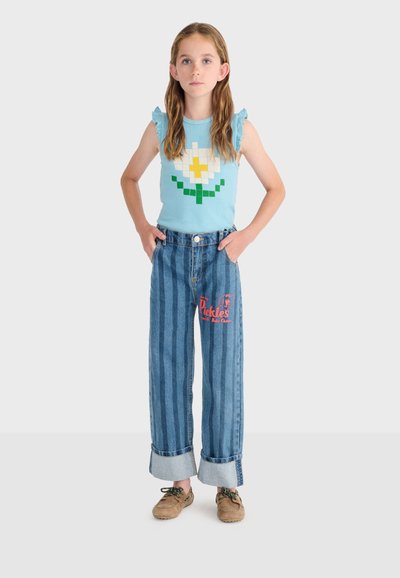 Fille debout les mains dans les poches, portant un haut bleu sans manches avec une marguerite pixellisée et un jean à jambes larges rayé avec un texte rouge.