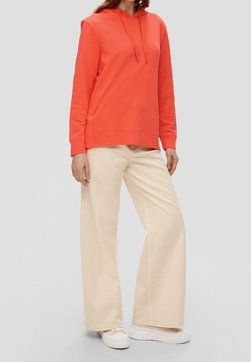 Korallfärgad huvtröja med långa ärmar och känguruficka, i kombination med off-white vida byxor och vita sneakers.