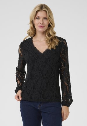 Haut à manches longues en dentelle noire avec un décolleté en V à volants et un motif floral. Le tissu est semi-transparent, associé à un jean noir.