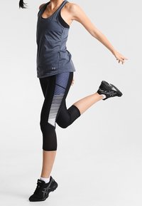 Sportovní tílko v tmavě šedé barvě, zkombinované s černými legínami s horizontálními pruhy. Model má na sobě černé běžecké boty.