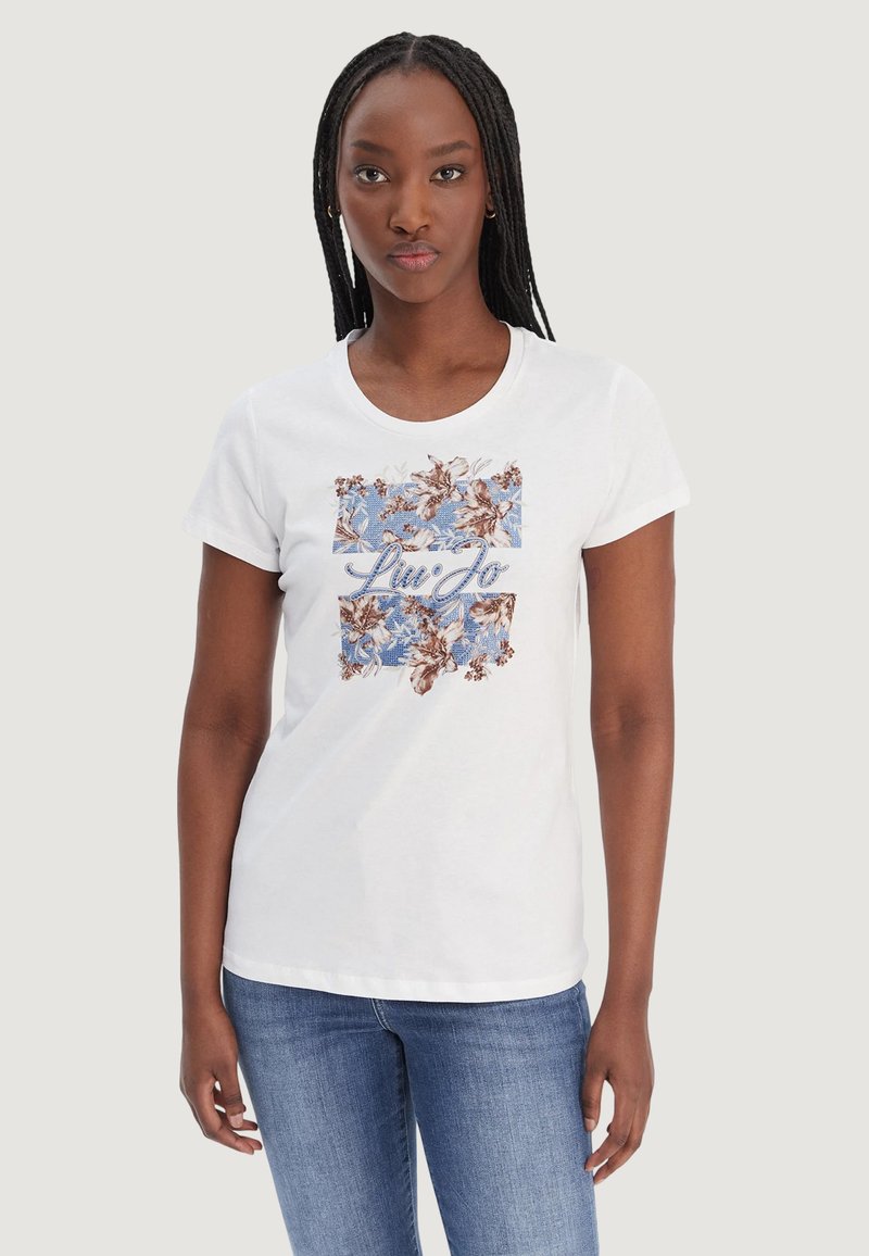 Femme aux cheveux tressés portant un T-shirt blanc à motif floral et un jean bleu, debout devant un fond clair uni.