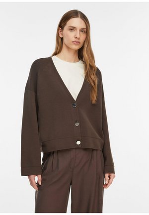 Femme aux longs cheveux portant un cardigan boutonné marron foncé par-dessus un haut blanc et un pantalon marron assorti et ample, sur fond blanc.