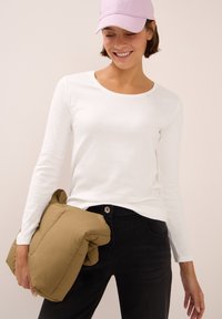 Femme souriante portant une chemise blanche à manches longues, un jean noir, une casquette rose, et tenant une veste marron repliée devant un fond uni.