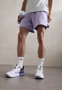 Witte sportieve schoenen met paarse accenten, lichtpaarse shorts en witte sokken met een zwart logo. Houdt dumbbells vast. Gelaagde achtergronden.