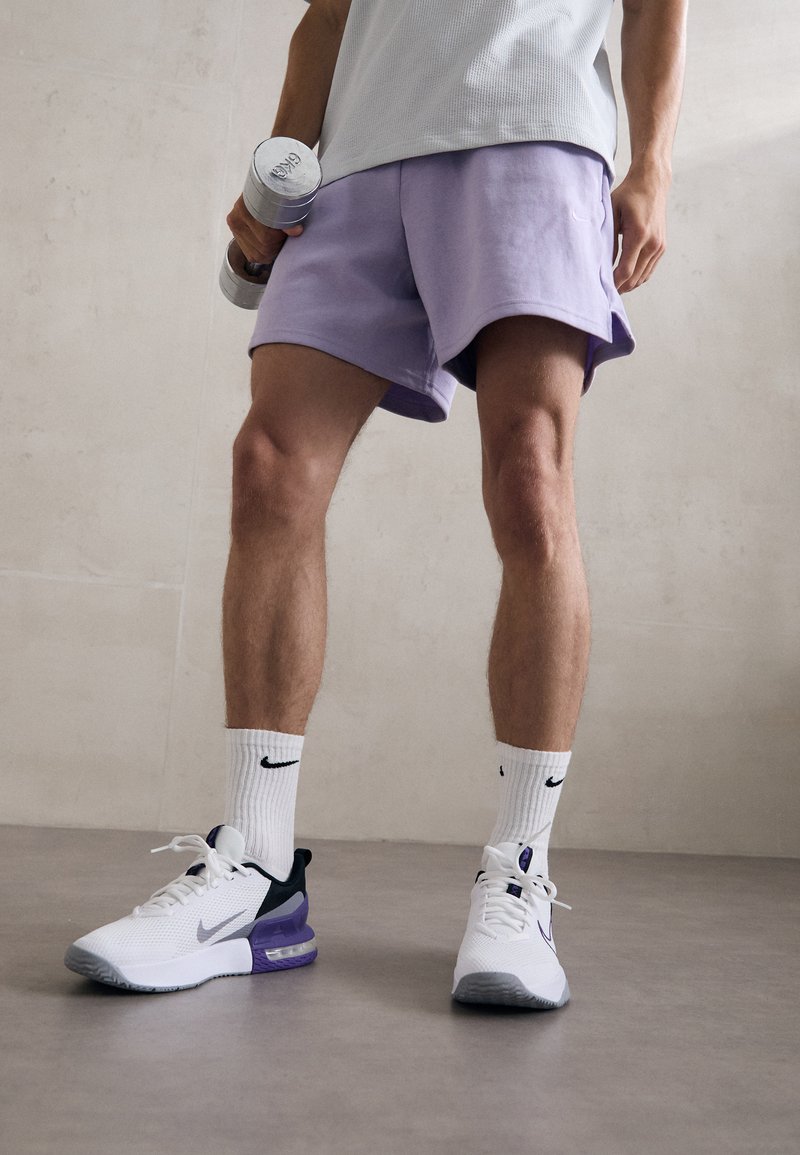 Witte sportieve schoenen met paarse accenten, lichtpaarse shorts en witte sokken met een zwart logo. Houdt dumbbells vast. Gelaagde achtergronden.