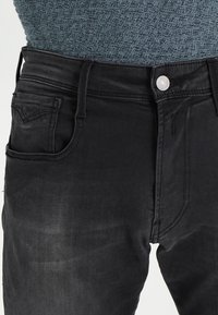 Vaqueros de denim negro con un diseño clásico de cinco bolsillos, que cuentan con un cierre frontal de botón, una textura sutil y una silueta ajustada.