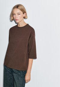 Woman with short blonde hair wearing a loose brown elbow-sleeve shirt and blue jeans, standing against a plain white background.