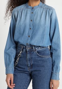 Chemise en denim bleu clair à boutons avec col mao, coutures froncées aux épaules et manches longues. Associée à un jean bleu taille haute.