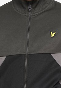 Chaqueta de cremallera que presenta una sección superior de color gris oscuro, una sección inferior negra y detalles en gris. Cuello acanalado y pequeño logotipo amarillo visible.