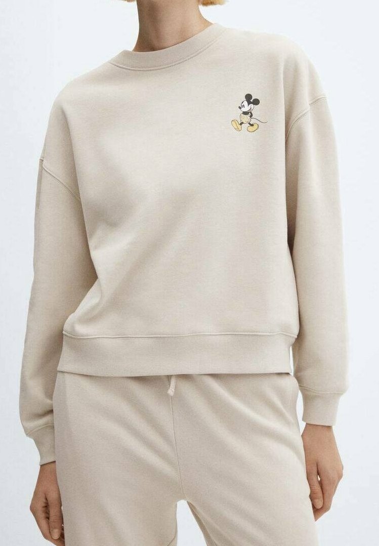 Sudadera beige con cuello redondo y puños de canalé, que presenta un pequeño bordado de Mickey Mouse en el lado izquierdo del pecho. Textura de tela suave.