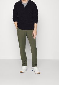 Dunkler Strickpullover mit Kragen, graues Rundhals-T-Shirt darunter, olivgrüne Hose und weiße Sneakers mit blauen Akzenten.