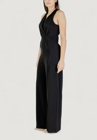 Donna in un tuta nera senza maniche, con gambe larghe e vita aderente, che indossa scarpe nude su uno sfondo chiaro.