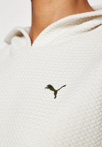 Bijela teksturirana dukserica s 3D uzorkom, s zelenim izvezenim Puma logotipom na prsima i kapuljačom sa vezicom.