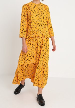 Woman wearing a yellow long-sleeve floral midi dress with ruffles paired with black oxford shoes, standing against a plain background.