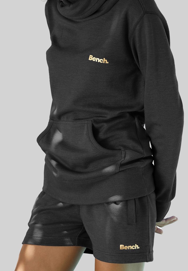 Personne portant un sweat à capuche noir Bench et un short assorti avec un logo Bench jaune sur la poitrine et la jambe gauche, debout devant un fond neutre.