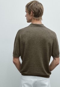 Hnědé pletené polo tričko s jemnou texturou, krátkými rukávy a malým límcem, nošené s bílými kalhotami, z pohledu zezadu.