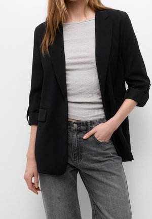 Femme portant un blazer noir aux manches retroussées, un haut côtelé gris clair et un jean noir délavé, main dans la poche sur fond blanc.