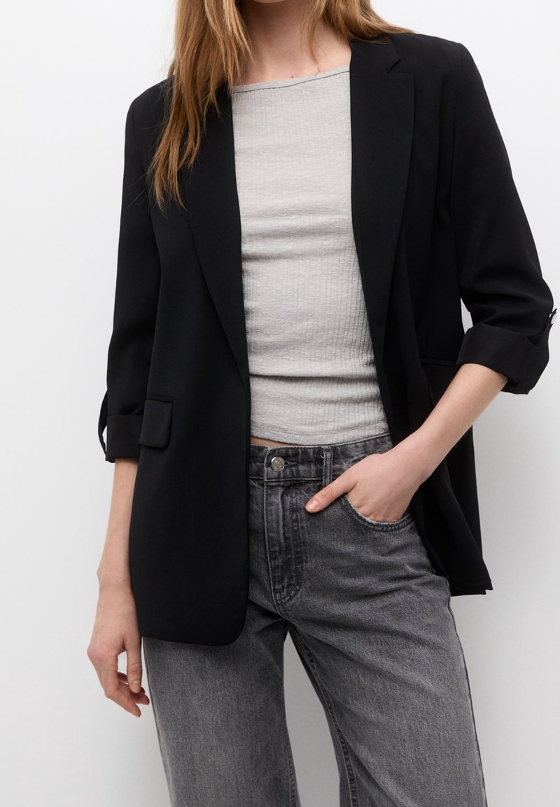 Femme portant un blazer noir aux manches retroussées, un haut côtelé gris clair et un jean noir délavé, main dans la poche sur fond blanc.