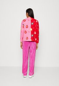 Le pull présente un design fendu avec des sections rose et rouge ; le côté rouge est orné de motifs colorés en forme de gouttes. Associé à un pantalon plissé rose.