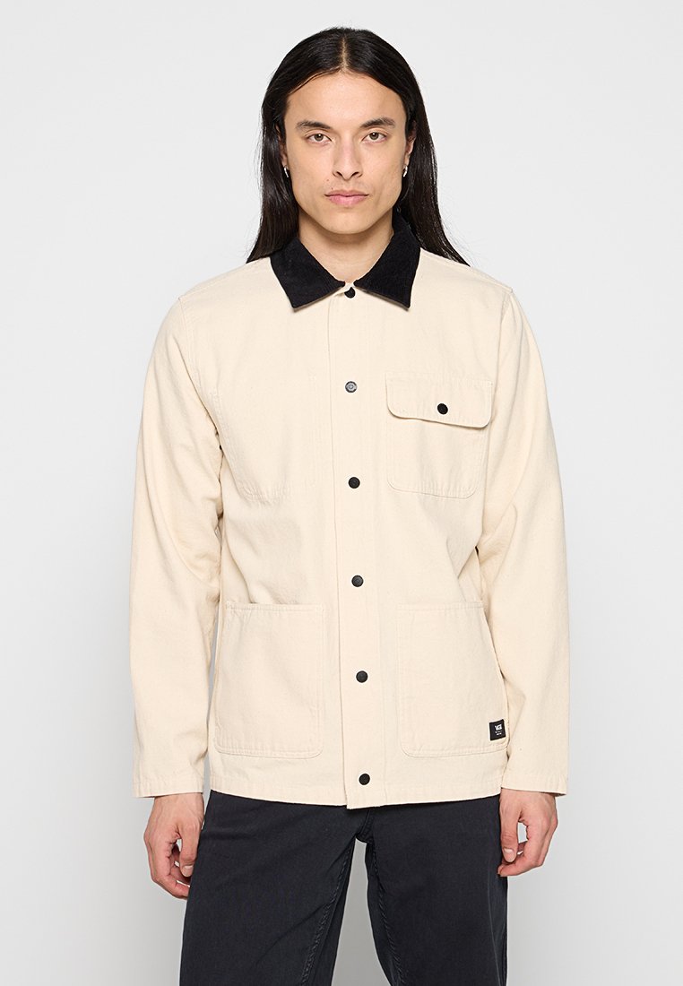 Chaqueta color beige claro con un cuello de pana negro, botones a presión en el frente, dos grandes bolsillos frontales y una pequeña etiqueta negra en el dobladillo.