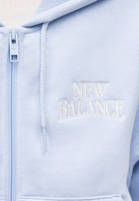 Ανοιχτό μπλε φούτερ με φερμουάρ, κατασκευασμένο από απαλό ύφασμα, με λευκό κεντητό λογότυπο "New Balance" στο αριστερό στήθος και υφασμένες κορδόνια.