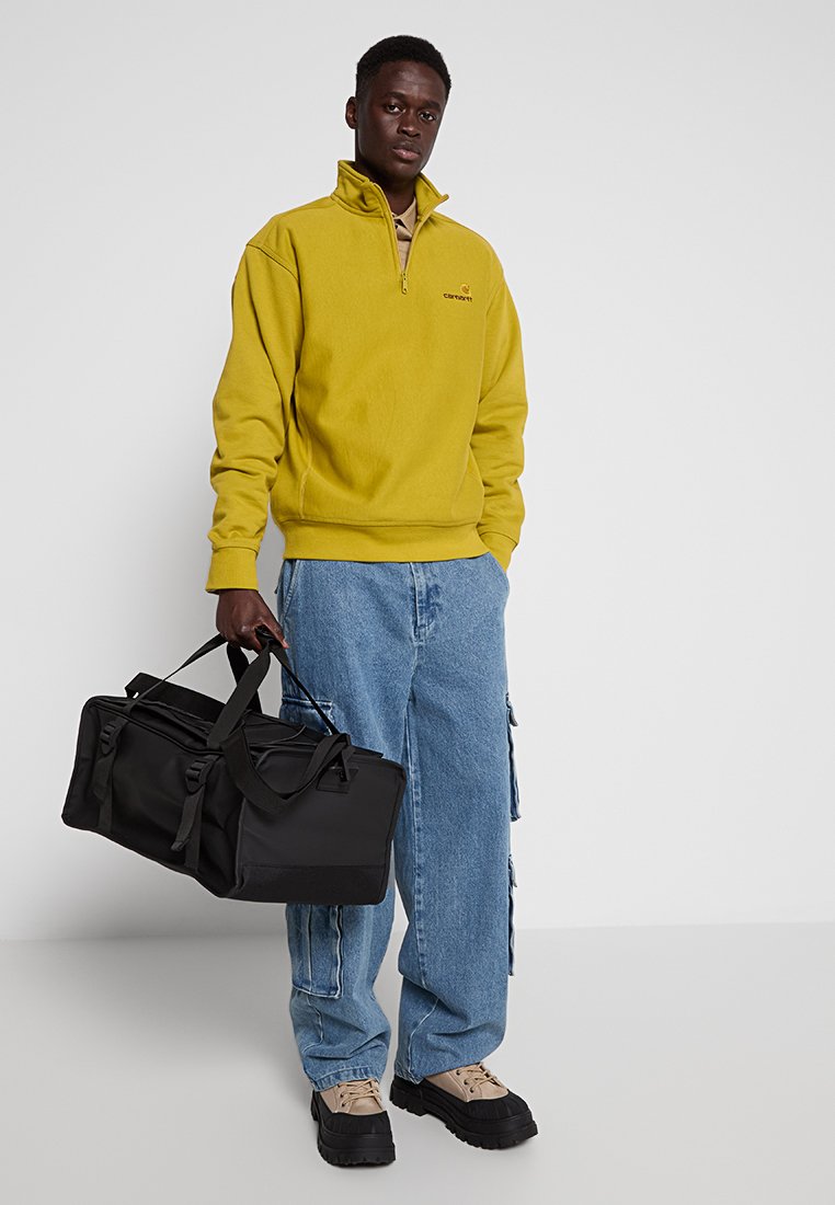 Pull jaune en polaire avec fermeture éclair quart, sac de voyage noir, jean bleu ample avec détails cargo, et baskets beiges avec une épaisse semelle noire.