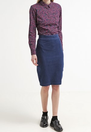 Vrouw draagt een paarse blouse met bloemenprint, een blauwe rok tot op de knie, zwarte sokken en zwarte schoenen met gespen, staand tegen een witte achtergrond.