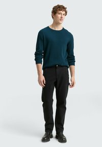 Teal geribbelde trui met lange mouwen, gecombineerd met zwarte broek. Afgemaakt met zwarte laarzen. Model staat tegen een lichte achtergrond.