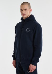 Tamnoplava zip-up dukserica od mekog materijala, s kengurujem džepom, kapuljačom s špagom i logotipom na prsima. Glatka tekstura.