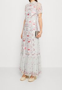 Maxiklänning i genomskinlig ljusgrå tyg med blommig broderi i rosa, lila och orange; volangärmar; lager på lager nederkant; matchad med en kuvertväska.