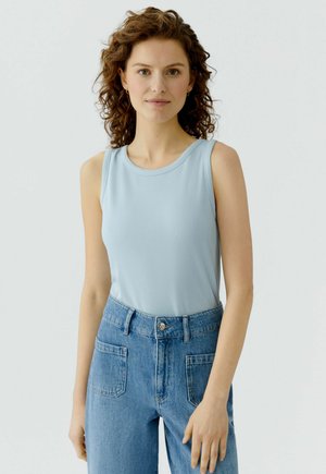 Jeune femme aux cheveux bouclés portant un débardeur côtelé bleu clair et un jean bleu taille haute, debout devant un fond clair uni.