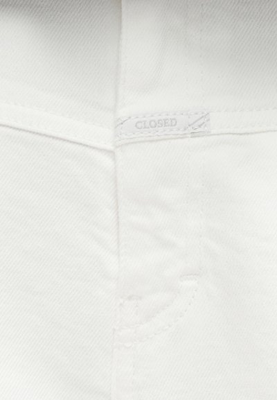 Λευκό ύφασμα τζιν με ορατές ραφές και μια μικρή κεντημένη ετικέτα με την επιγραφή "CLOSED" κοντά στην περιοχή της τσέπης.