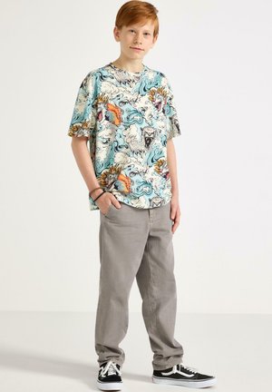 Rudovlasý chlapec v barevném tričku s potiskem draka a mraků, šedých kalhotách a černých teniskách, stojící s rukama v kapsách na jednobarevném pozadí.