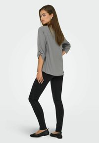 Mujer con cabello largo castaño que lleva una blusa gris de manga 3/4, jeans ajustados negros y zapatos planos negros, de pie con una mano en la cadera mirando hacia atrás.
