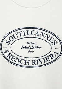 Valkoisessa hupparissa on sininen soikio grafiikka. Teksteissä lukee "SOUTH CANNES," "FRENCH RIVIERA," ja hotellitiedot perinteisellä fonttisuunnittelulla.
