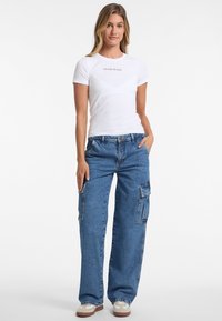 Weißes T-Shirt mit kurzen Ärmeln und in Schwarz bedrucktem "Guess Jeans", kombiniert mit lässigen, hellblauen Denim-Cargohosen mit Seitentaschen.