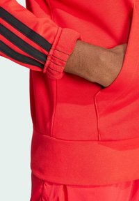 Felpa sportiva rossa con strisce nere sulle maniche, caratterizzata da polsini e vita a coste, texture liscia e una vestibilità comoda.