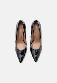 O pereche de pantofi cu toc înalt, lucioși, de culoare neagră, cu vârful ascuțit, având içuri de culoare bej și branding "Anna Field", văzuți de sus pe un fundal alb.