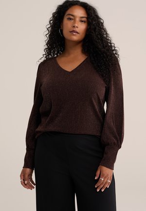 Femme aux longs cheveux bouclés portant un haut à manches longues col en V marron foncé chatoyant et un pantalon noir, debout devant un fond beige.