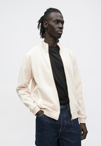 Lysebeige bomberjakke med geometrisk tekstur, hvite detaljer og glidelås. Brukt over en svart t-skjorte, kombinert med mørk denim.