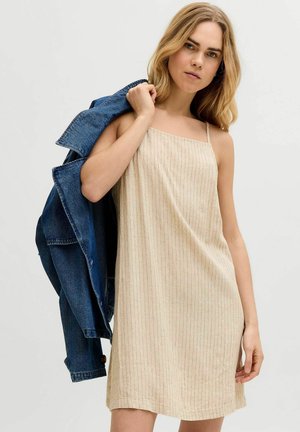 Jeune femme portant une robe beige sans manches à fines rayures, tenant une veste en jean bleue sur l'épaule, sur un fond uni.